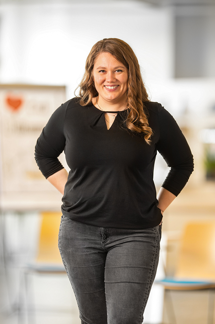 Eine Frau mit langen gewellten Haaren, die ein schwarzes Oberteil und dunkle Jeans trägt, steht in einem Raum und lächelt mit den Händen hinter dem Rücken. Im Hintergrund sind Stühle und ein Schild zu sehen, die leicht verschwommen sind.