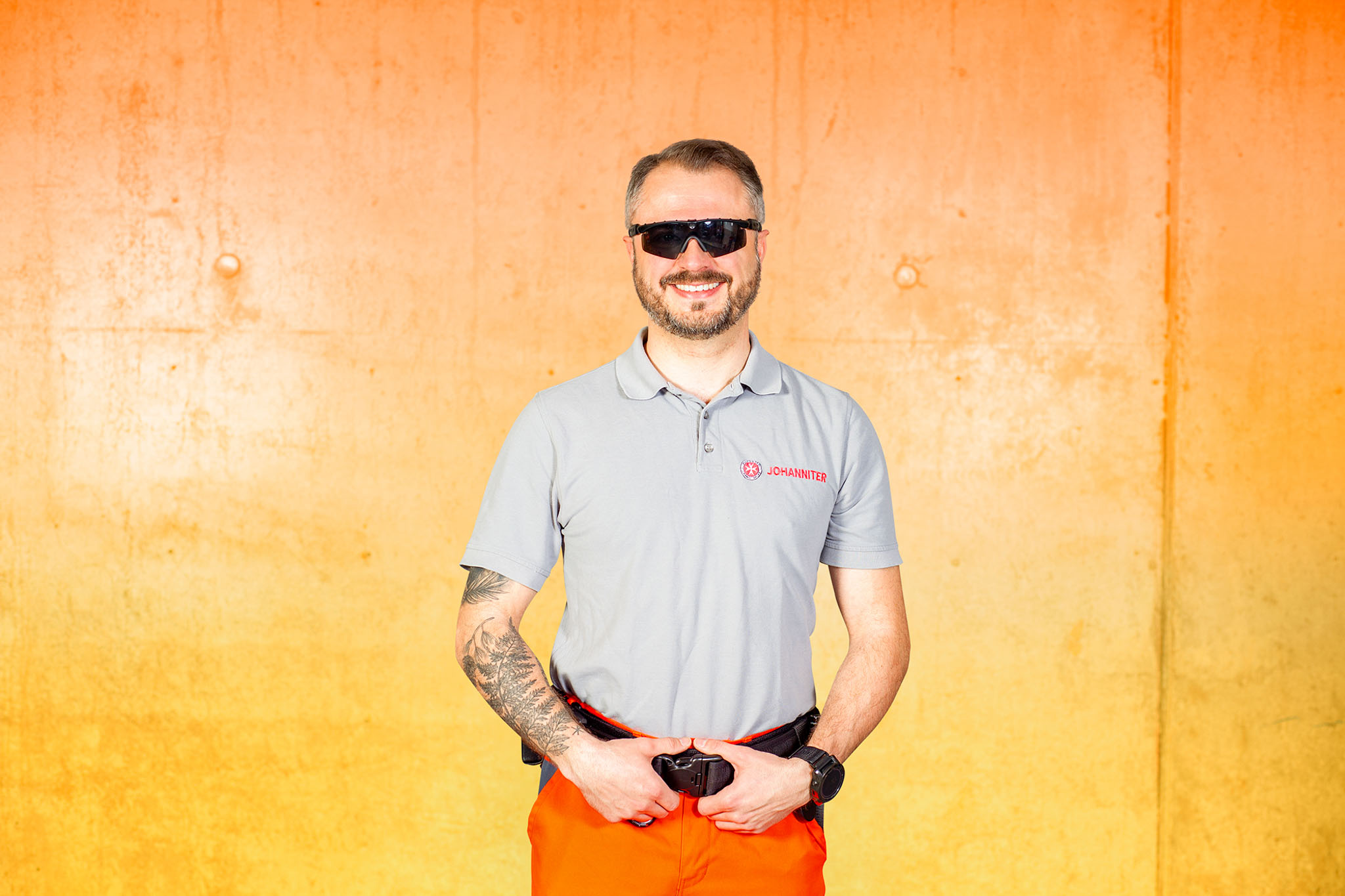 Ein Mann mit Bart, Sonnenbrille und einem Tattoo auf dem linken Arm steht lächelnd vor einer orangefarbenen Wand. Er trägt ein hellgraues Poloshirt, eine orangefarbene Hose und eine schwarze Uhr. Seine Hände ruhen auf seinem Gürtel.