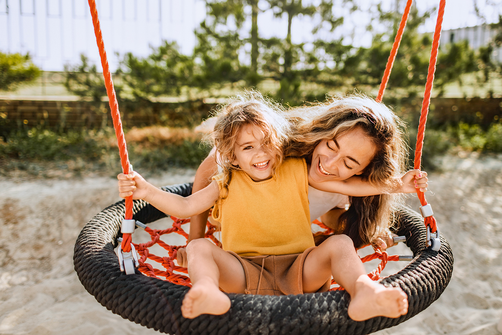 Eine Frau und ein junges Mädchen lächeln und lachen zusammen, während sie an einem sonnigen Tag auf einer runden Seilschaukel auf einem Spielplatz sitzen. Das Mädchen trägt ein gelbes T-Shirt und Shorts, und die Frau umarmt sie von hinten. Im Hintergrund sind Bäume und Sand zu sehen.