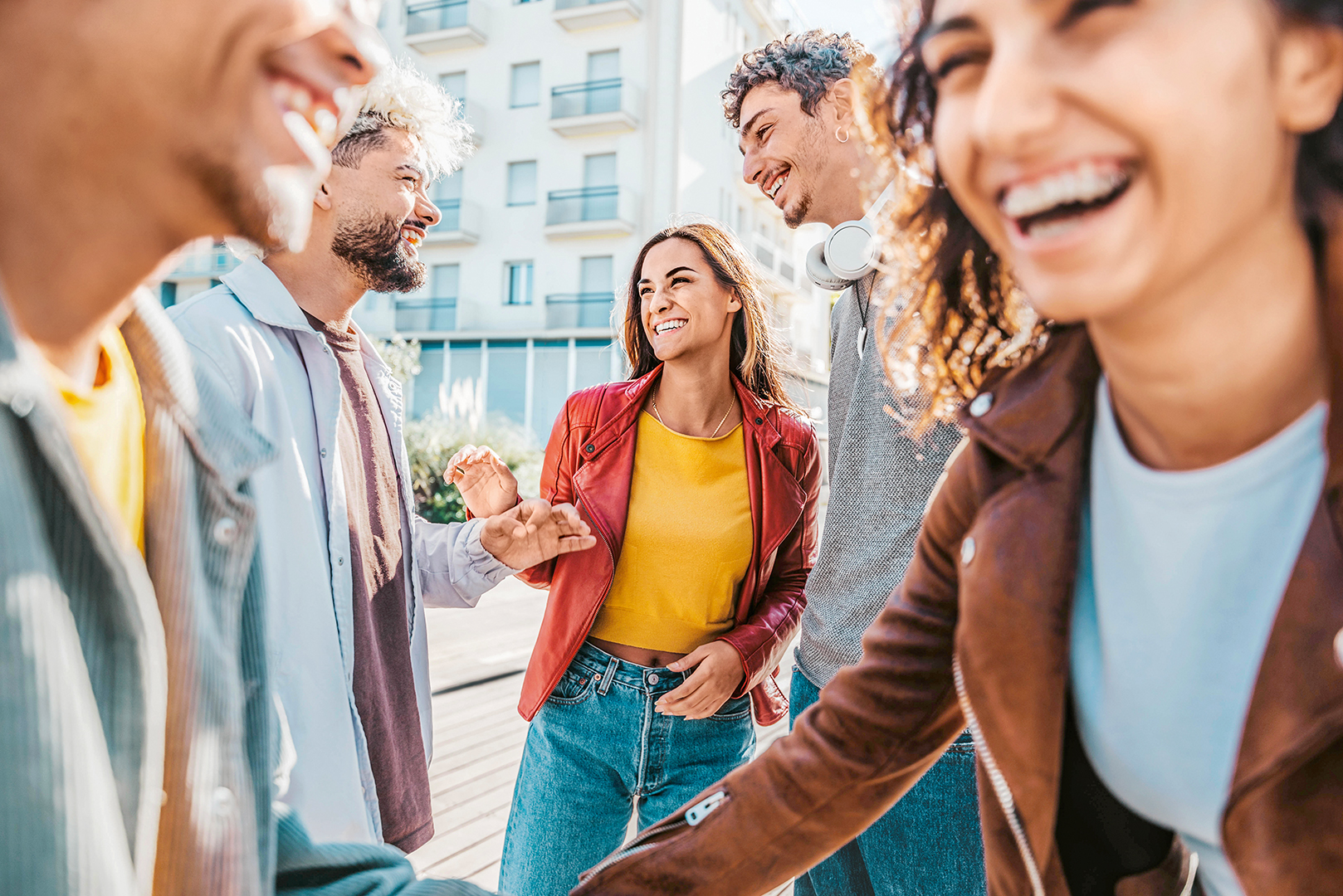 Eine Gruppe von vier jungen Erwachsenen steht an einem sonnigen Tag im Freien, lächelt und lacht zusammen. Sie tragen legere Kleidung und scheinen die Gesellschaft des anderen in einer städtischen Umgebung zu genießen.