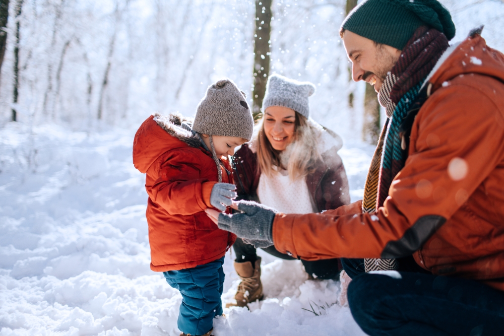 Eine Familie genießt einen verschneiten Tag im Freien. Ein Kind in einem roten Mantel und einer grauen Mütze spielt im Schnee, während zwei Erwachsene, warm gekleidet und lächelnd, glücklich zuschauen. Im Hintergrund sind schneebedeckte Bäume zu sehen, und um sie herum fallen sanft Schneeflocken.