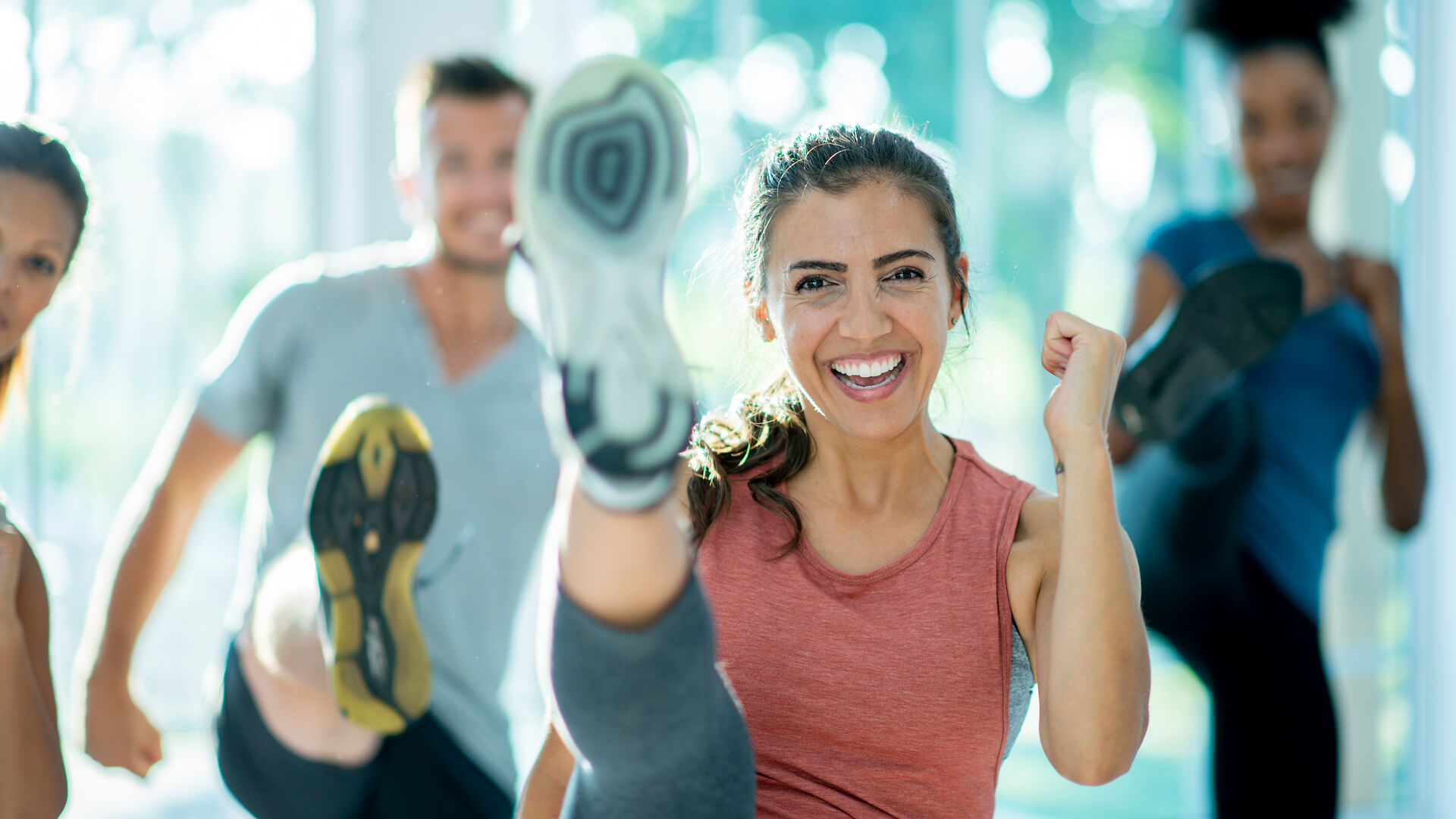 Eine Gruppe von Menschen treibt drinnen Sport, lächelt und schwingt die Beine hoch. Im Mittelpunkt steht eine Frau mit einem ärmellosen Oberteil, die energiegeladen und fröhlich aussieht, während die anderen im Hintergrund verschwommen sind.